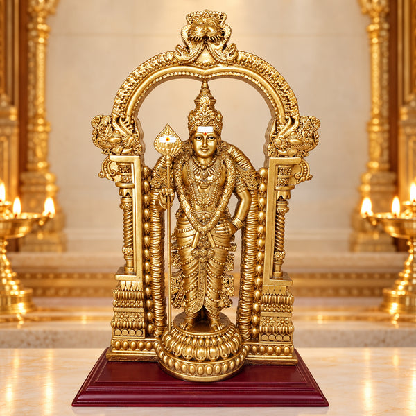 Gold Murugan statue with temple arch and vel on a wooden base, placed in a pooja room with brass lamps